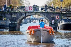 Een rondvaartboot vaart onder een brug door in Amsterdam, met een schipper in een blauwe KINboat-shirt die de route navigeert, terwijl mensen op de brug wandelen en fietsen
