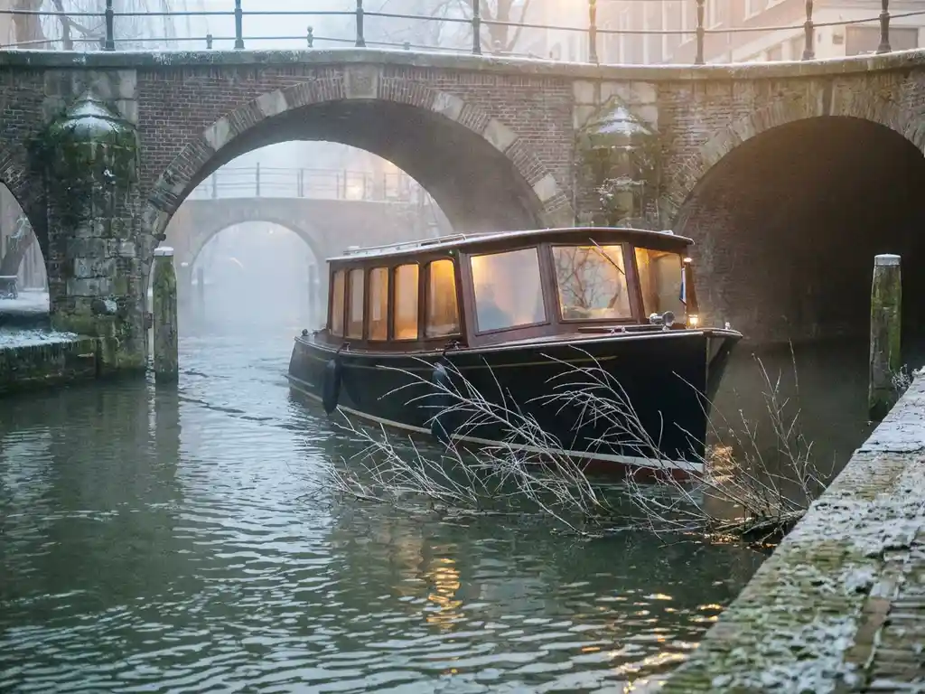 Electric canal boat gliding through a misty Utrecht canal in winter, frost-dusted stone bridge overhead, amber cabin light glowing warmly.