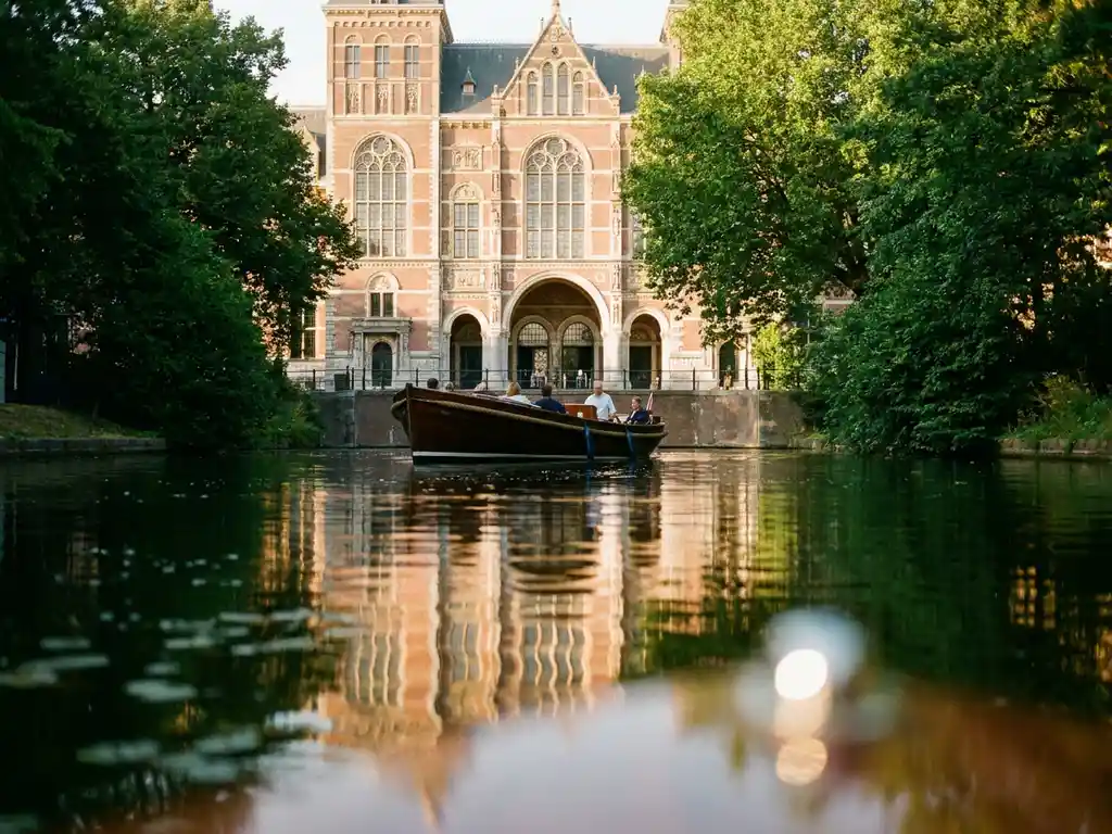 Electric canal boat gliding past the Rijksmuseum's ornate brick facade, reflected in still green water under warm golden afternoon light.
