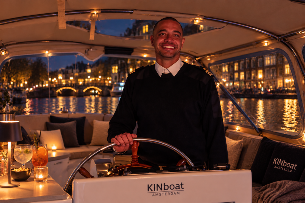 Lachende schipper van KINboat aan het roer van een overdekte sloep op de Amsterdamse grachten